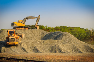 Yellow wheel loader and excavator are working in quarry against the background of crushed stone storage.