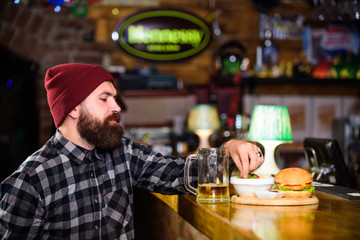 Enjoy meal in pub. High calorie snack. Brutal hipster bearded man sit at bar counter. Hipster relaxing at pub. Pub is relaxing place to have drink and relax. Man with beard drink beer eat burger menu