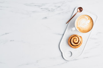 Freshly baked cinnamon roll with spices and cocoa filling and coffee or cappuccino with latte art on white serving plate over white marble background. Top view. Copy space for text. Swedish breakfast.