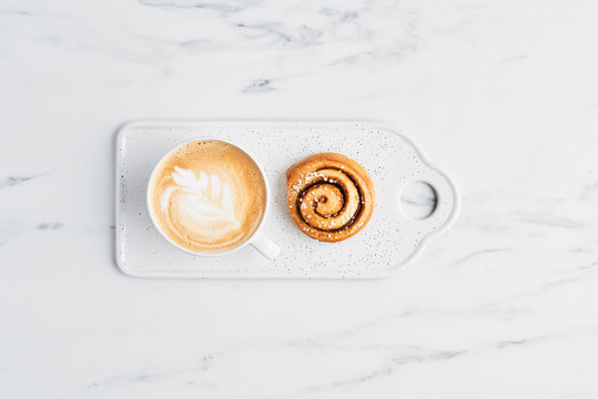 Freshly baked cinnamon bun with spices and cocoa filling and coffee or cappuccino with latte art on white serving plate over white marble background. Top view. Copy space for text. Swedish breakfast.
