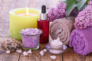 Red bottle with aromatic oil, burning candle, bowls with sea salt, lilac flowers and towels.