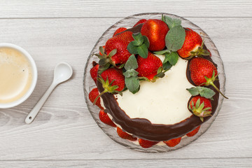 Homemade chocolate cake decorated with fresh strawberries on glass plate and cup of coffee on gray background.