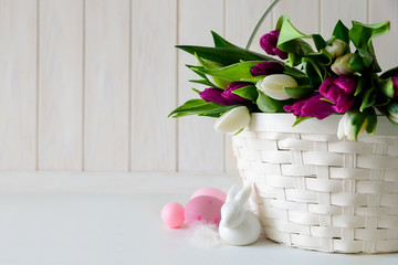 Basket with white and purple tulips, Easter eggs and rabbit on white wooden background. Copy space