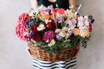 Very nice young woman holding beautiful wicker basket full of blossoming hydrangea, peonies, eustoma, mattiola, carnations, roses, pistachio leaves in different colours on the grey wall background