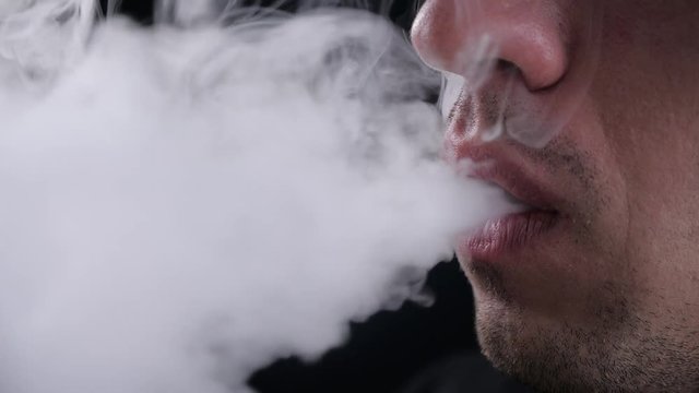 Close-up of man vaper exhaling big clouds of smoke with e-cigarette vape on black background in slow motion