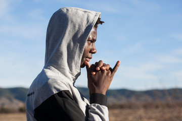 chico africano con sudadera gris y capucha puesta en un entorno natural con cielo azul y día...