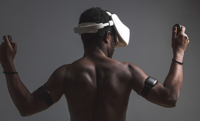 Strong African boxer with bare chest wearing mobile virtual reality headset prepares to fight with virtual opponent, standing isolayed against dark wall. Sport and augmented virtual reality concept