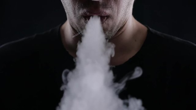 Close-up of man vaper exhaling big clouds of smoke with e-cigarette vape on black background in slow motion