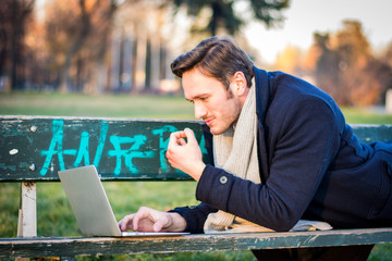 Handsome elegant businessman sitting on a wooden bench working outdoors in an urban park typing...