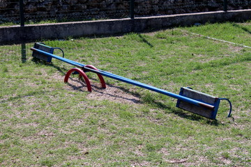 Broken vintage retro old blue metal playground seesaw with wooden seats surrounded with grass and metal fence in background on warm sunny day