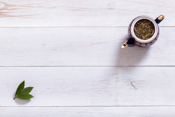 Teapot and tea time leaves set on white wooden table, black herbal homemade hot beverage in porcelain pot with green leaf, english tea ceremony background, top view from above, copy space