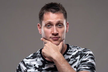 Portrait of expressive uncertain man looking at camera with hands on chin over gray background. 