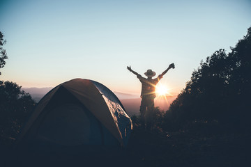 Happy hiker man in a hat standing holding a coffee cup near camping tent on mountains at sunset background. travel concept.