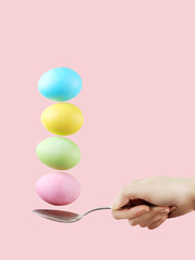 Female hand holds a spoon on which multi-colored eggs are balanced, on a pink background. Unusual design, Easter concept, copy space.