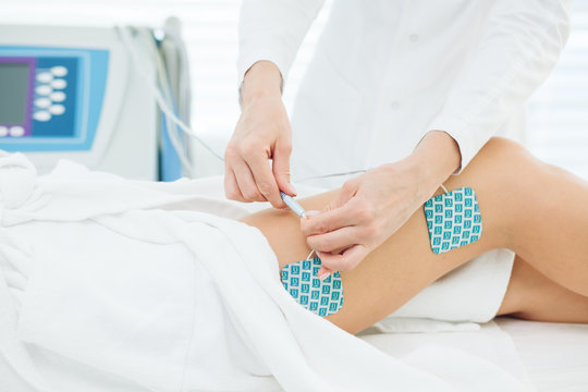 Beautician hands operating with eElectric device, sticking electrodes to female hips. Myostimulation session performed on the slim female legs with healthy skin