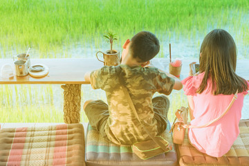 Back view of brother and sister are playing mobile phone games and relaxing with beverage at the cottage in the rice fields.