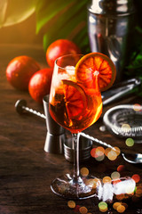 Obraz na płótnie Canvas Aperol spritz cocktail in big wine glass with bloody oranges, summer Italian fresh alcohol cold drink. Wooden bar counter background with tools, summer mood concept with palm trees, selective focus