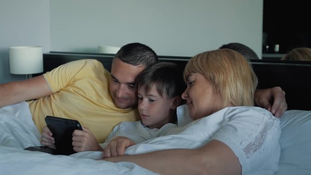 A happy family with one child lying on the bed and using a digital tablet and smartphones. Modern technologies of virtual life