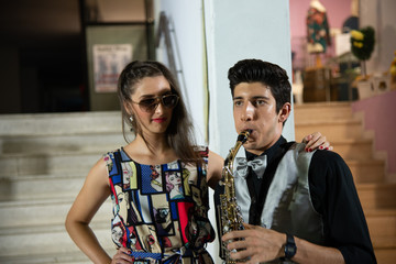 Couple of young people in love. The boy plays the saxophone, the girl puts a hand on the boy's shoulder. The brown-haired male in gray waistcoat rings, the girl with glasses in long fancy dress.