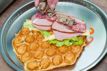 Bubble waffle, process of making waffles with tomatoes, lettuce, cheese, red sweet peppers, smoked meat, salami sausage and champignon mushrooms. Fast food concept, fast food, restaurant