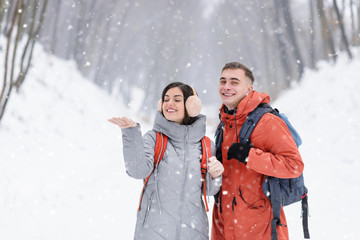 Brunette girl catches a snowflake on hand and blonde boy rejoices the snow and smiling to the camera while walking in the snowy forest with backpacks.