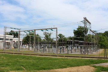 Fototapeta na wymiar Industrial power plant with multiple electrical power transformers connected with glass and ceramic insulators to dense wires surrounded with metal fence and high uncut grass with trees and cloudy blu