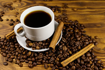 Cup of coffee, roasted coffee beans and cinnamon sticks on wooden table