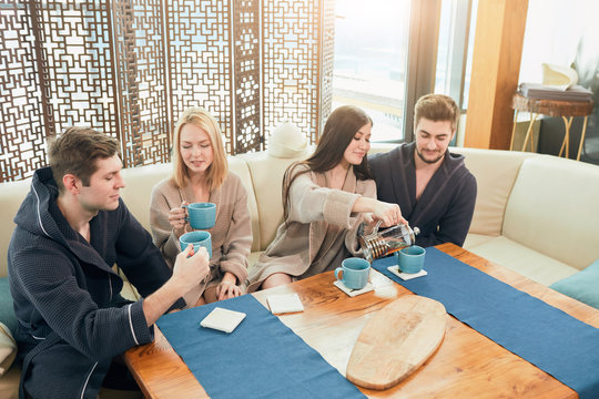 Cheerful young male and female friends in soft bathrobes sitting at the table with cups ,chilling in spa after massage procedure