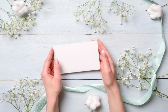 Beautiful female hands holding empty paper card with blank space on light blue wooden desk with flowers. Tender greeting card composition for womens or mothers day, easter, spring holidays. - Powered by Adobe