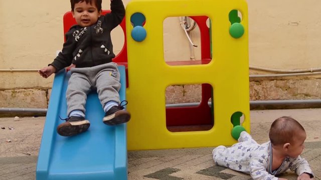 siblings playing in the backyard, toddler sliding down from plastic play house while his baby brother is having tummy time