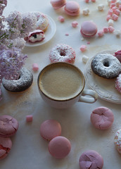 Cup of cappuccino, fresh colorful donuts, strawberry macarons and bouquet of lilac on white marble table