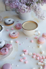 Cup of cappuccino, fresh colorful donuts, strawberry macarons and bouquet of lilac on white marble table