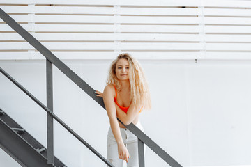 Fit and sporty blonde girl posing in a stylish white loft on a yellow background. Sports, fitness, diet, and healthy lifestyle concept. Positive woman with curly hair in orange top and white pants