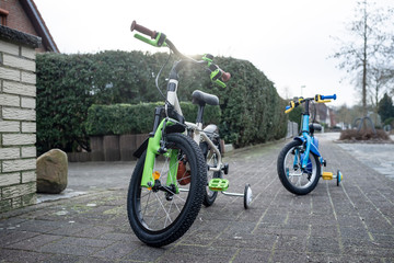 Two children's bicycles stand on the sidewalk in a provincial European city, in the background you can see the yard with a hedge and part of the house.