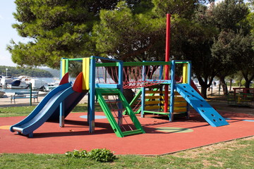 Multifunctional outdoor public playground equipment with blue plastic slide next to climbing net and wall connected with safety net tunnel on local playground surrounded with grass and trees with sea 