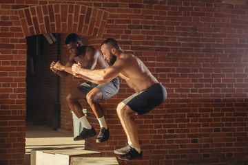 Obraz premium Two multi ethnic male sportsmen are jumping on plyo box, close up. African fitness instructor with his caucasian trainee doing box jump workout at cross fit gym