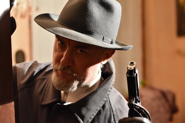 Closeup portrait of a mature caucasian  man in a fedora and raincoat (investigator, private detective or hitman) holding a gun. Interior scene selective focus