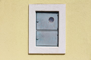Outdoor locked metal electrical meter box with switch box mounted on house wall with new facade surrounded with white frame on warm sunny day