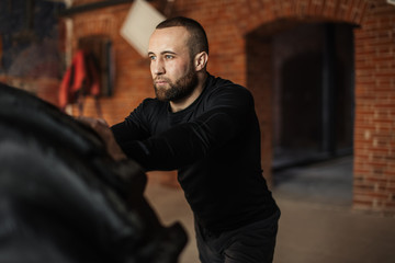 Obraz premium Silny sportowiec przewracający ogromną oponę w studiu CrossFit z minimalistycznym wnętrzem ceglanych ścian. Jest to ćwiczenie, które symuluje jednocześnie martwy ciąg, gwałtowne unoszenie się z kolan i parcie w górę.