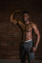 Handsome African American bodybuilder man, with naked muscular torso showing musculs against red brick wall.