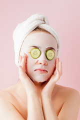 Beautiful young woman is applying a cosmetic tissue mask on a face with cucumber on a pink background