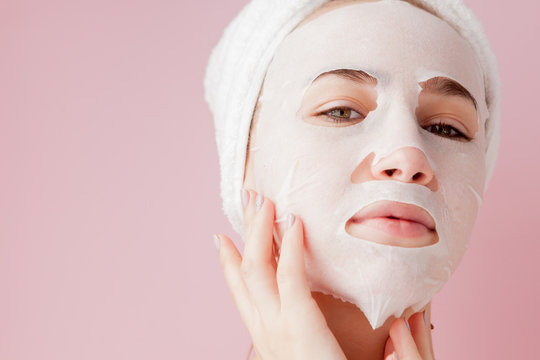 Beautiful young woman is applying a cosmetic tissue mask on a face on a pink background
