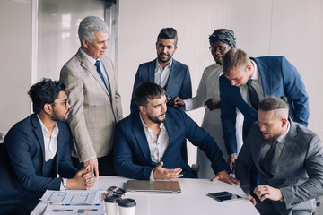Confident team of age and race diverse male professionals dressed in formal wear excited with success of construction company gatther around boss tabledesk.