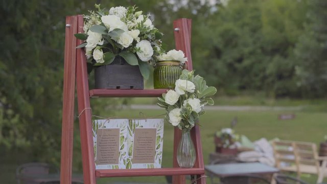 On a wooden stepladder there is a glass vase with white roses, a black wooden box with a bouquet of white flowers, signs with a list of guests and numbering of tables, shooting on the street on a summ