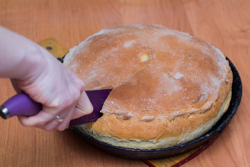 A woman cuts a meat pie with a knife. A traditional home-made pastry dish lies in a black pan on the table. © PAVEL GERASIMENKO