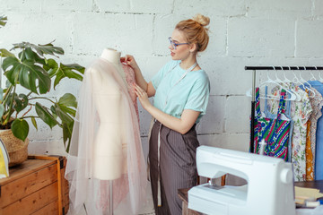 Skillful fashion designer stands in a dressmaker studio near the mannequin and fixes lace fabric on...