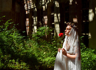 A young woman prays inside a large ruin.  She is dressed in white and wears  a white veil and a thorn wreath on her head. 