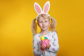 Obraz na płótnie Canvas Cute adorable Caucasian blonde girl in white dress wearing pink Easter bunny ears holding small basket with eggs in studio on yellow background. Funny kid child celebrating Christian holiday