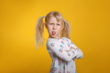 Grumpy angry Caucasian blonde girl with blue eyes in white dress posing in studio on yellow background with arms crossed on chest. Kid child expressing negative emotion showing tongue