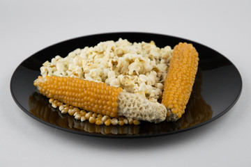 Making popcorn at home in microwave: Well-dried corn on the cob are among grains and the finished product in black plate, white, side view